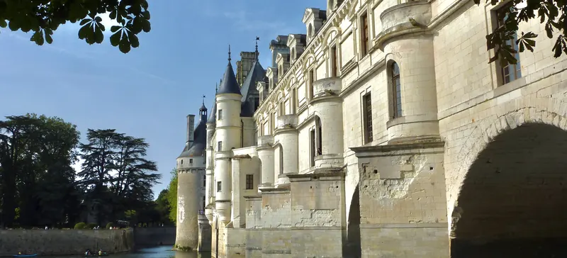 La façade du château de Chenonceau vue d’en bas depuis le Cher, ses arches de pierre, ciel bleu.