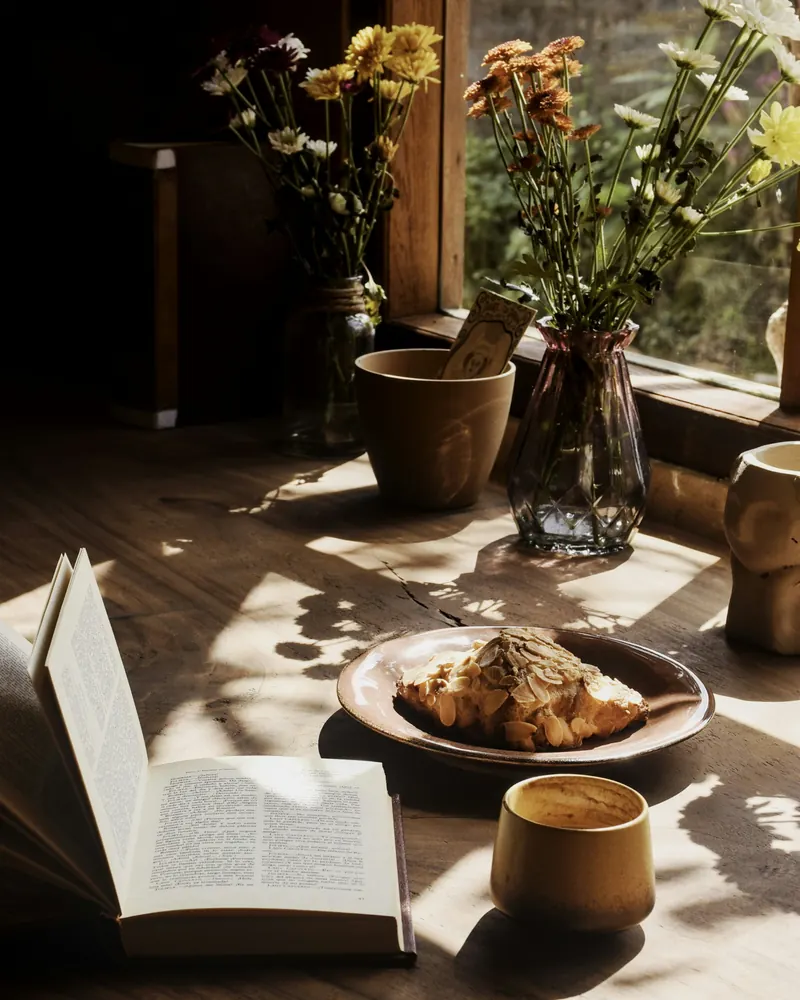 Une pâtisserie aux amandes et un café posés sur une table en bois, lumière du matin, fleurs dans des vases près de la fenêtre.