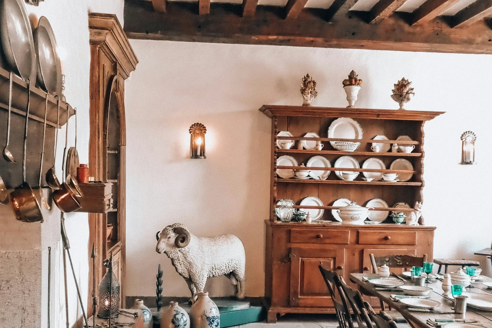 Salle à manger aux poutres apparentes, vaisselier en bois chargé d’assiettes blanches, appliques allumées, casseroles en cuivre suspendues.